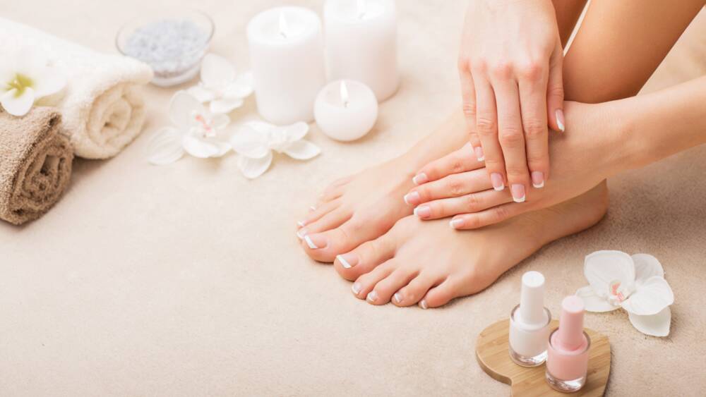 Zwei gepflegte Hände liegen gefaltet auf gepflegten Füßen. Die Nägel sind frisch lackiert. Im Hintergrund sind Kerzen, Nagellacke, Handtücher und Blüten von Blumen zu sehen.