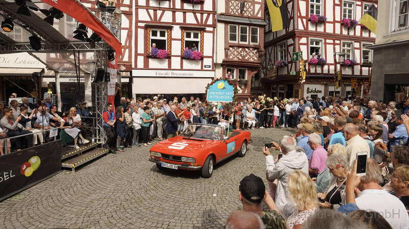 Ein roter Oldtimer mit der Moselweinkönigin fährt über den von zahlreichen Besuchern gefüllten Marktplatz. Auf der Bühne sitzen Ehrengäste.