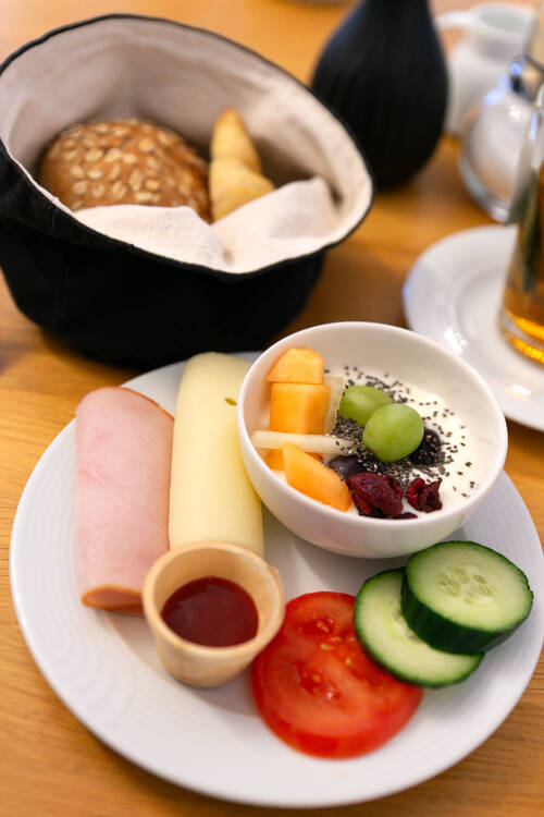 Ein Teller mit einer Auswahl an Belägen, Obst und Gemüse vom Frühstücksbuffet. Neben dem Teller steht ein kleines Körbchen mit frischen Croissants und Brötchen.