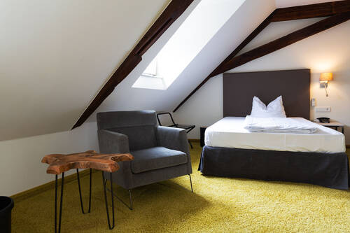 Ein großes Einzelbett steht in einem kleinen, gemütlichen Raum mit knallig grünem Teppichboden. Aus der Dachschräge und der Decke ragen alte Holzbalken hervor.