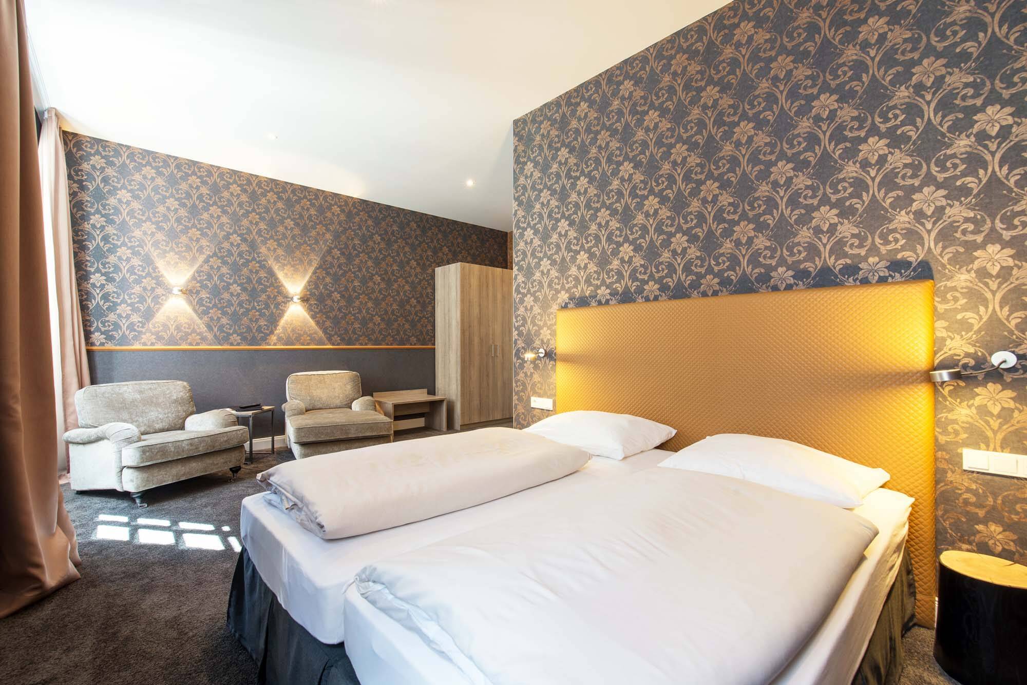 Ein Doppelbett mit goldenem Kopfteil steht in einem Zimmer mit auffälliger, grauer Tapete mit goldenem Muster. Die Lampen über den Nachttischen spenden gemütliches Licht. Im hinteren Teil des Raums stehen zwei graue Sessel und ein Beistelltisch als gemütliche Sitzecke.