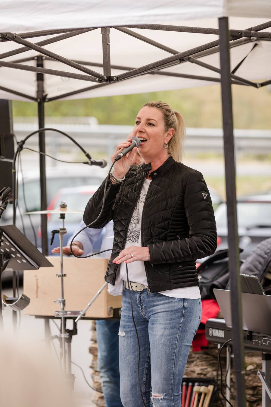 Eine blonde Frau singt in ein Mikrofon. Hinter ihr steht ein E-Piano und zwei weitere Bandmitglieder