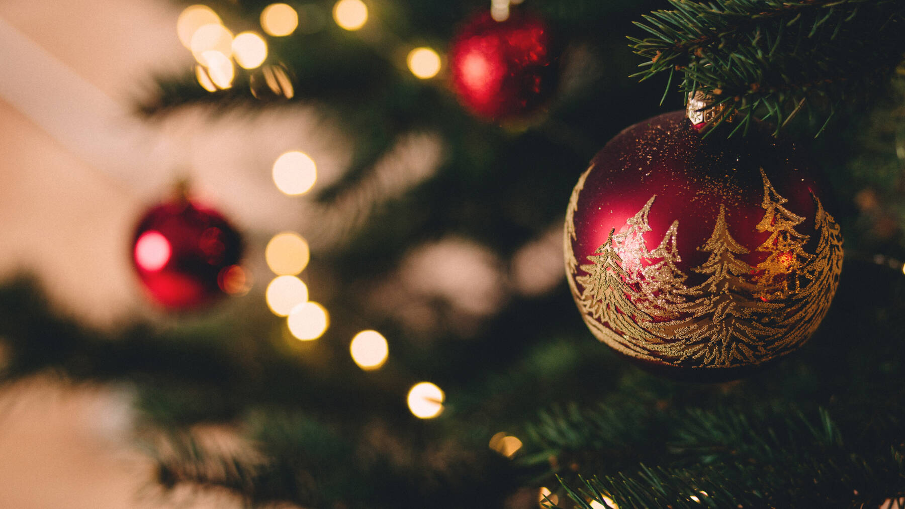 Im Fokus steht eine rote Christbaumkugel mit goldenen Tannenbäumen als Verzierung. Im unscharfen Hintergrund leuchten die Lichterkette des Weihnachtsbaums und weitere rote Kugeln.