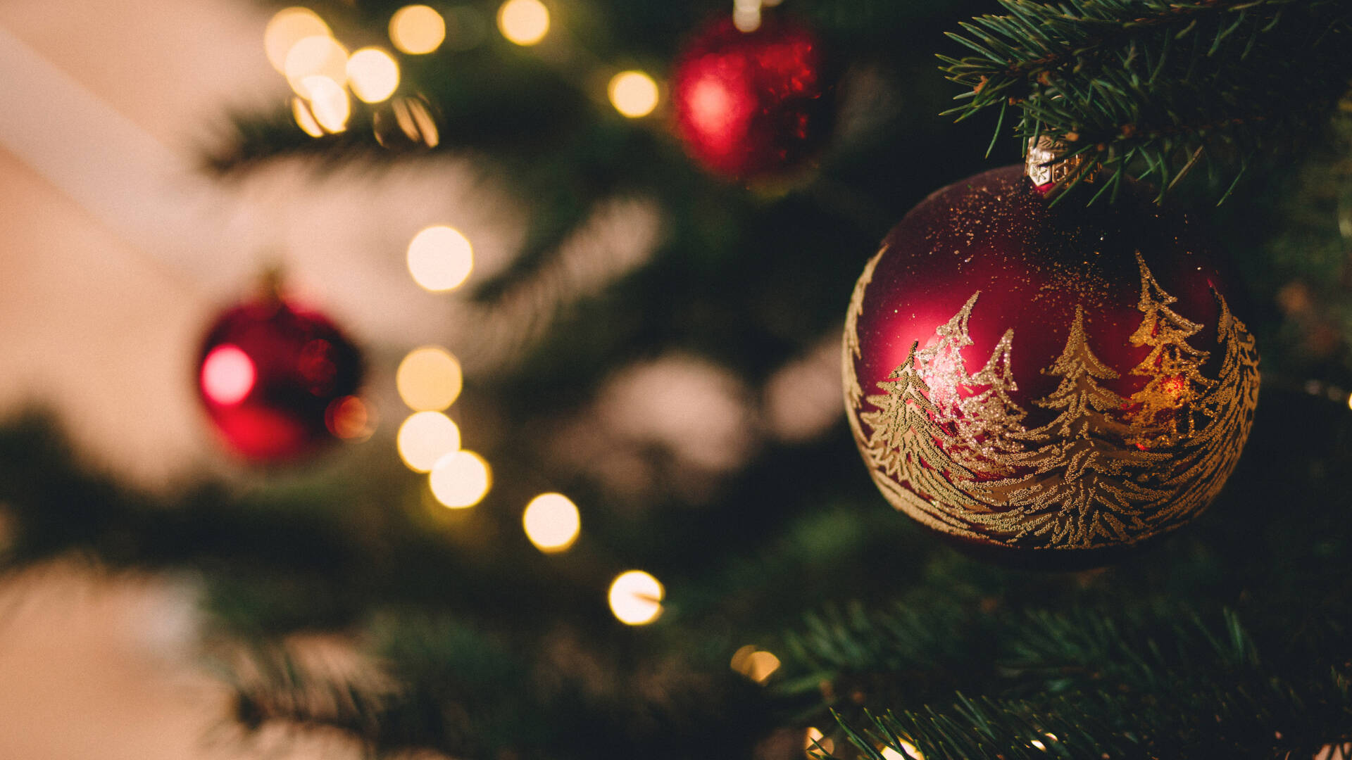 Im Fokus steht eine rote Christbaumkugel mit goldenen Tannenbäumen als Verzierung. Im unscharfen Hintergrund leuchten die Lichterkette des Weihnachtsbaums und weitere rote Kugeln.