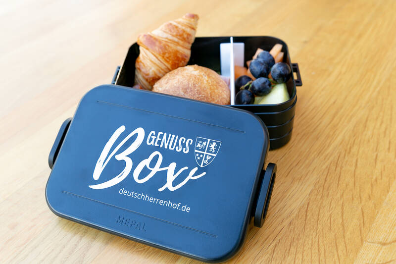Eine Brotdose steht auf dem Tisch. Der schwarze Deckel trägt die Aufschrift ‚Genussbox‘ und liegt angelehnt an der Dose. In der Dose befinden sich verschiedene Früchte, ein Brötchen und ein Croissant.