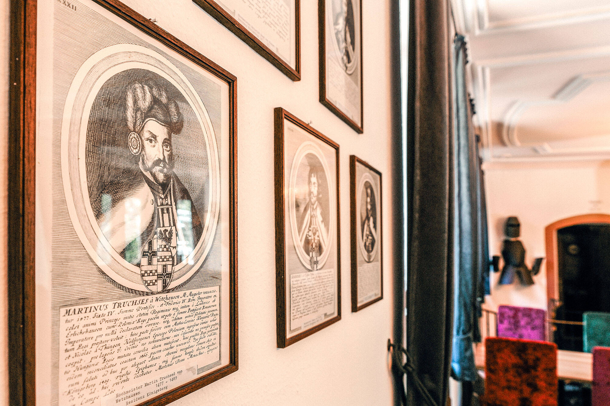 Detailaufnahme der Dekoration im Rittersaal. An der Wand hängen Strichzeichnungen historischer Persönlichkeiten des Hauses, darunter ein Hochmeister. Im unscharfen Hintergrund ist eine Ritterrüstung zu sehen, die an der Wand zur Dekoration hängt.