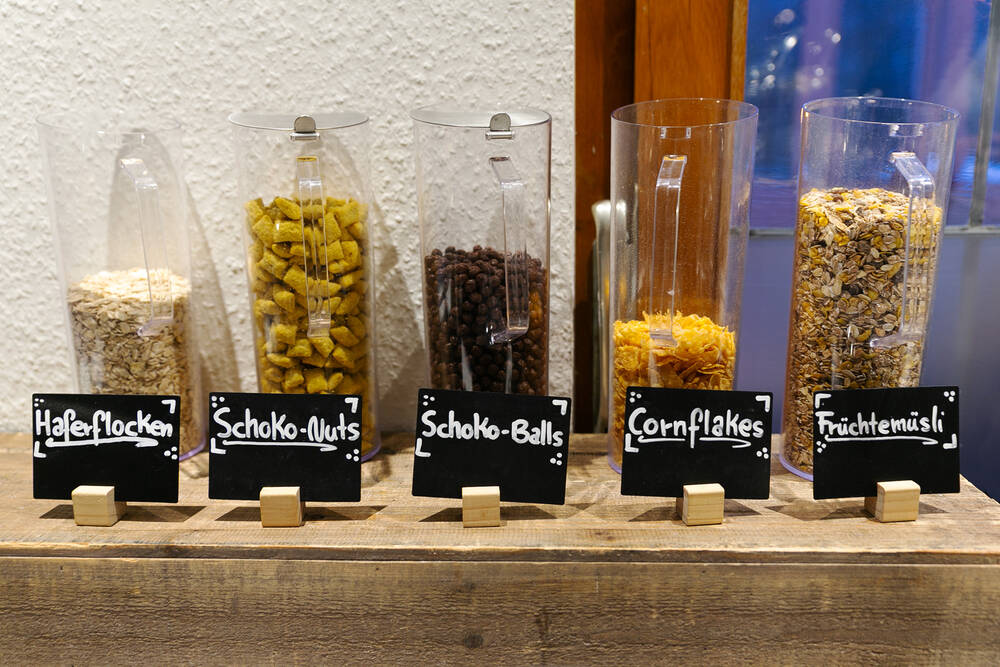Auf dem Frühstücksbuffet sind fünf Gläser mit verschiedenen Müsli- und Cornflake-Sorten aufgestellt. Jedes Glas ist mit einem kleinen Schieferschild versehen, auf dem die jeweilige Sorte angegeben ist.