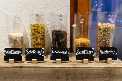 Auf dem Frühstücksbuffet sind fünf Gläser mit verschiedenen Müsli- und Cornflake-Sorten aufgestellt. Jedes Glas ist mit einem kleinen Schieferschild versehen, auf dem die jeweilige Sorte angegeben ist.