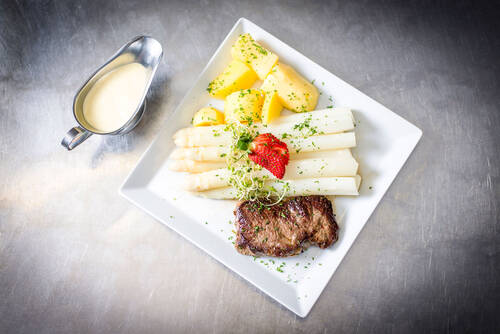 Auf einem quadratischen Teller liegen frisch gekochte Stangen weißer Spargel, dazu ein saftiges Stück Fleisch und Kartoffeln. Daneben steht eine Sauciere mit Sauce Hollandaise.