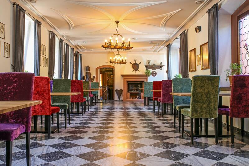 Blick in den Rittersaal des Deutschherrenhofs. Der Boden ist mit weißen und schwarzen Fliesen kariert. Rechts und links stehen bunte Stühle an Tischen. An der Decke hängen Kronleuchter, die mittig in einem Stuckrahmen platziert sind. Die Fenster sind von bodenlangen Vorhängen eingerahmt.