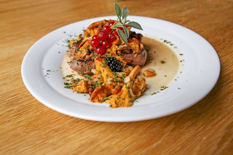 Ein Stück Fleisch liegt auf einem Teller, serviert mit Pfifferlingen, Soße, Preiselbeeren und Brombeeren.