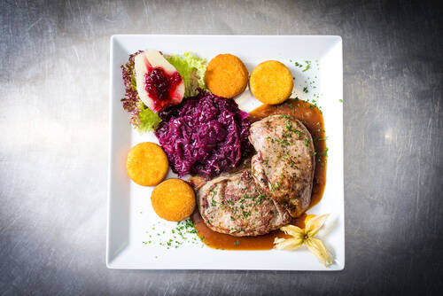 Auf einem quadratischen Teller liegt ein Stück Wild mit Rotkohl und mehreren Camembert-Talern. Eine halb aufgeschnittene Birne mit Preiselbeeren und eine Physalis dienen als Dekoration.
