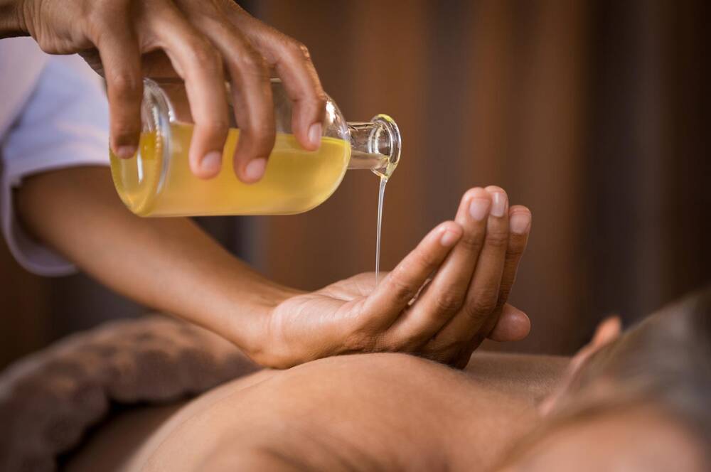 Eine Person liegt auf dem Bauch. Eine andere Person gießt Massageöl aus einer Glasflasche in ihre Handfläche, um es aufzutragen.