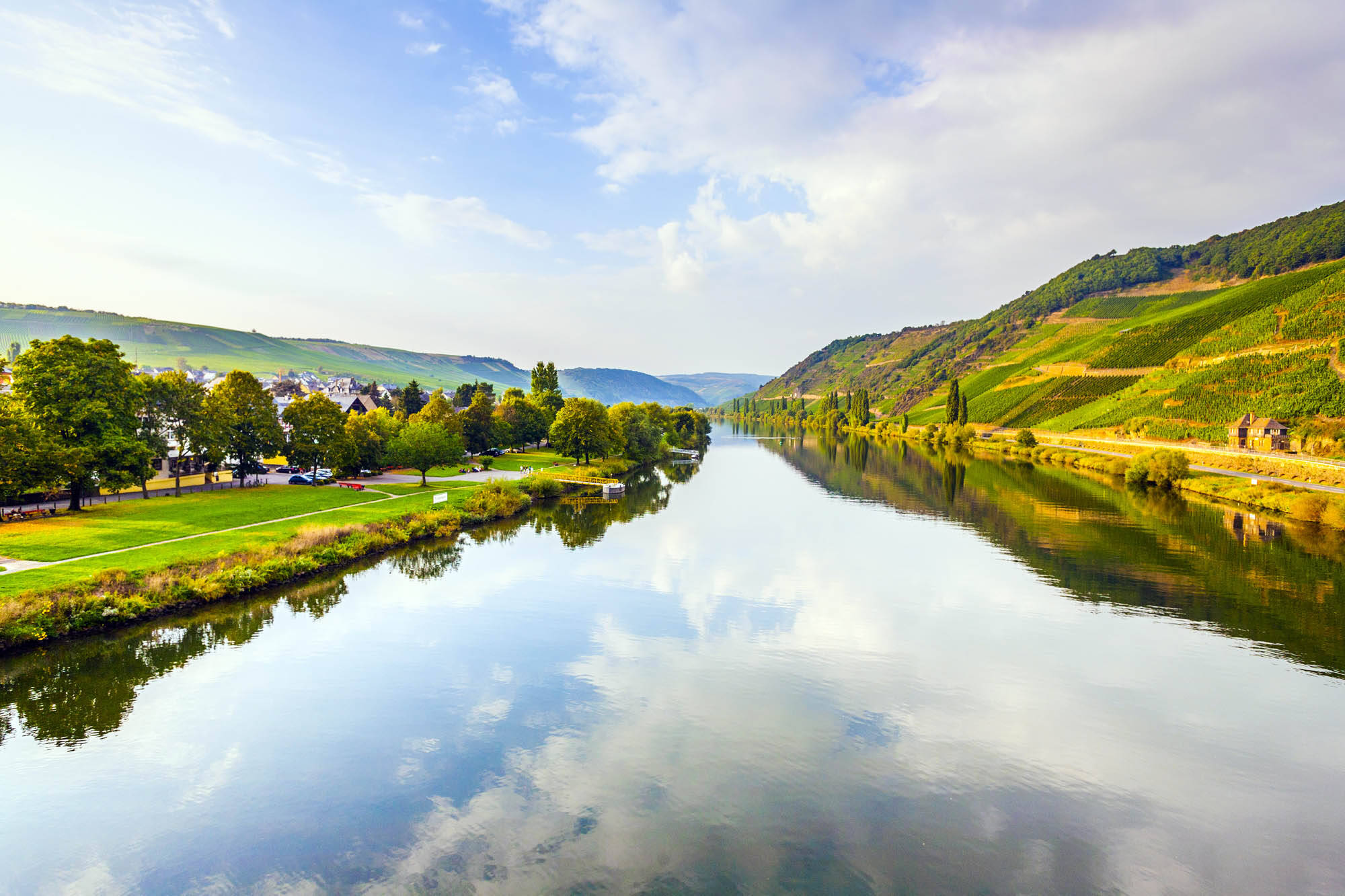 Das spiegelglatte Wasser der Mosel reflektiert den strahlend blauen Himmel. An den Uferseiten erstrecken sich Weinberge, Felder und Bäume.