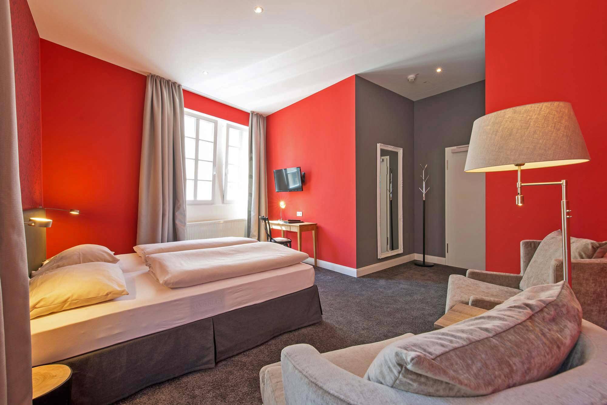 Blick in ein Doppelzimmer im Hotel Deutschherrenhof. Die Wände sind knallrot gestrichen, der Teppichboden ist grau. Beigefarbene Möbel, ein Doppelbett, ein kleiner Schreibtisch sowie eine Sitzecke mit zwei Sesseln und einer Lampe gehören zur Ausstattung.