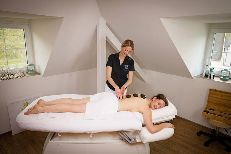 Eine Frau liegt mit einem Handtuch um die Hüfte auf einer Massageliege und erhält eine Hot-Stone-Massage von einer Mitarbeiterin.