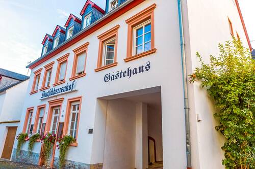 Die Front des Gästehauses. Auf dem weißen Haus mit terracottafarbenen Fenstereinfassungen steht in großen Buchstaben "Deutschherrenhof Gästehaus". Auf den Fensterbänken des Erdgeschosses sind Blumenkästen mit blühenden Pflanzen angebracht.