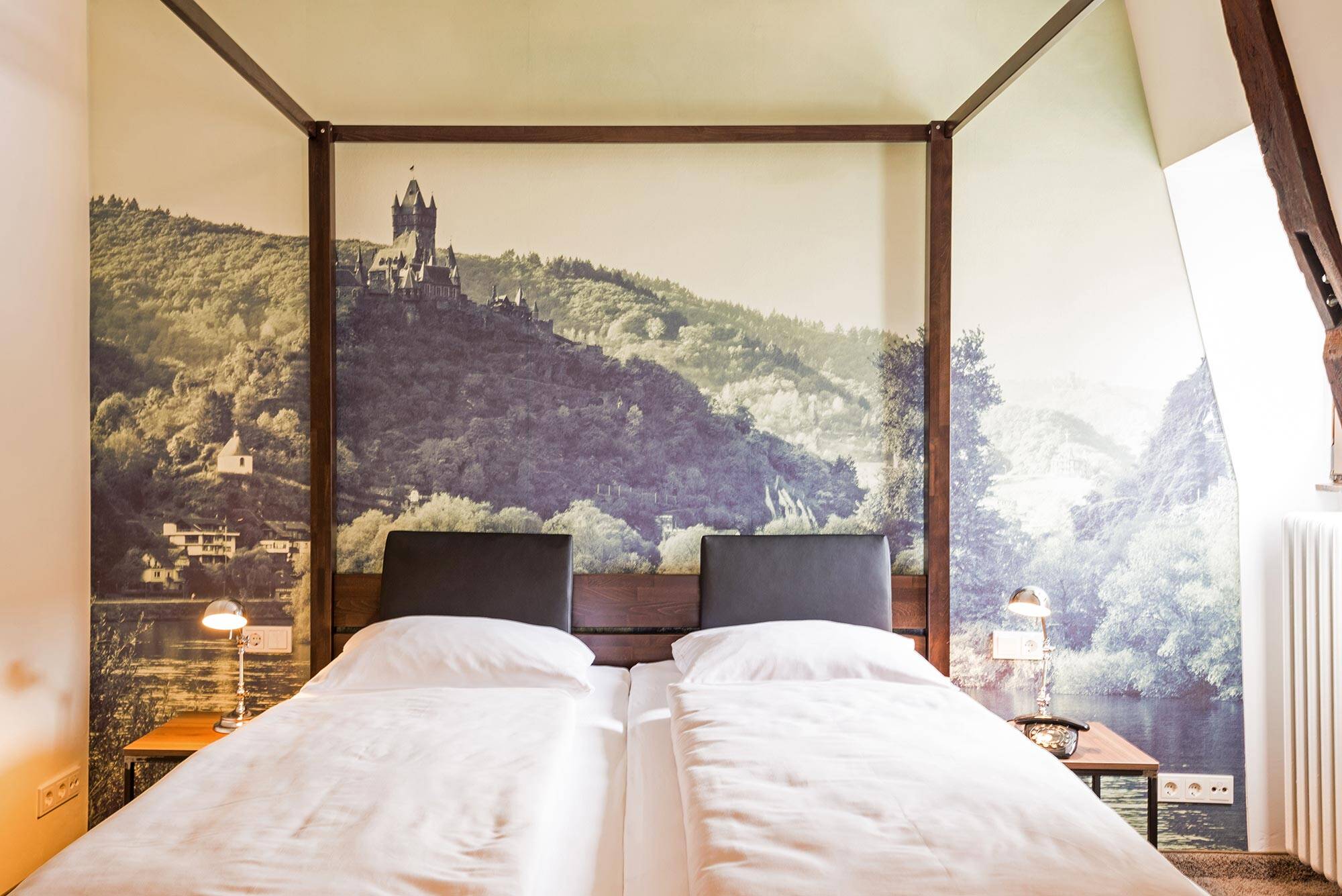 Ein Himmelbett aus dunklem Holz steht vor einer Wand mit einem großformatigen Schwarz-Weiß-Foto der Burg Cochem.