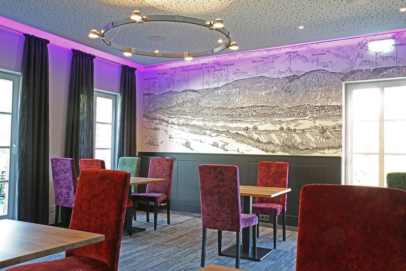 In der Speisekammer des Deutschherrenhofs stehen bunte Stühle an kleinen Tischen. Die Wand am Kopfende zeigt eine Zeichnung der Moselregion. Tageslicht strömt durch Fenster, die von bodenlangen Vorhängen umhüllt sind, und erhellt den Raum.