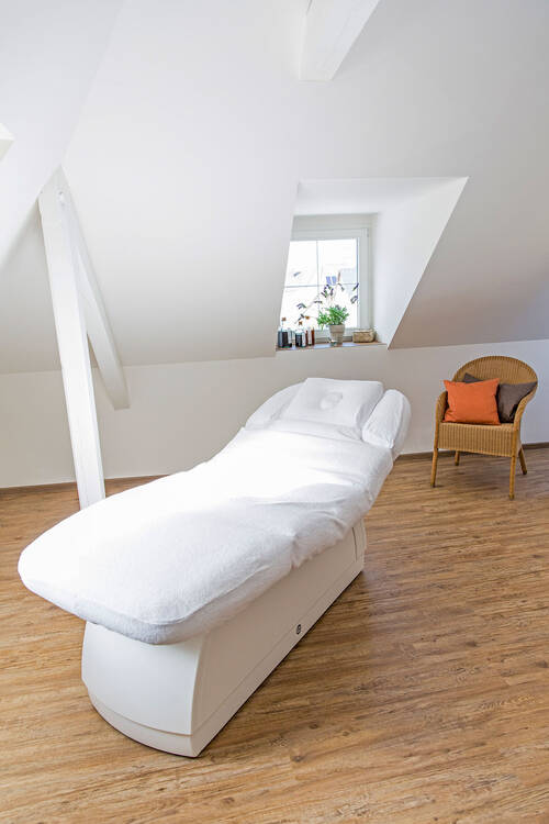 Die weiße Kosmetik- und Massageliege steht in einem großen, weiß gestrichenen Raum. Der helle Holzboden verstärkt die helle, freundliche Atmosphäre des Raums.
