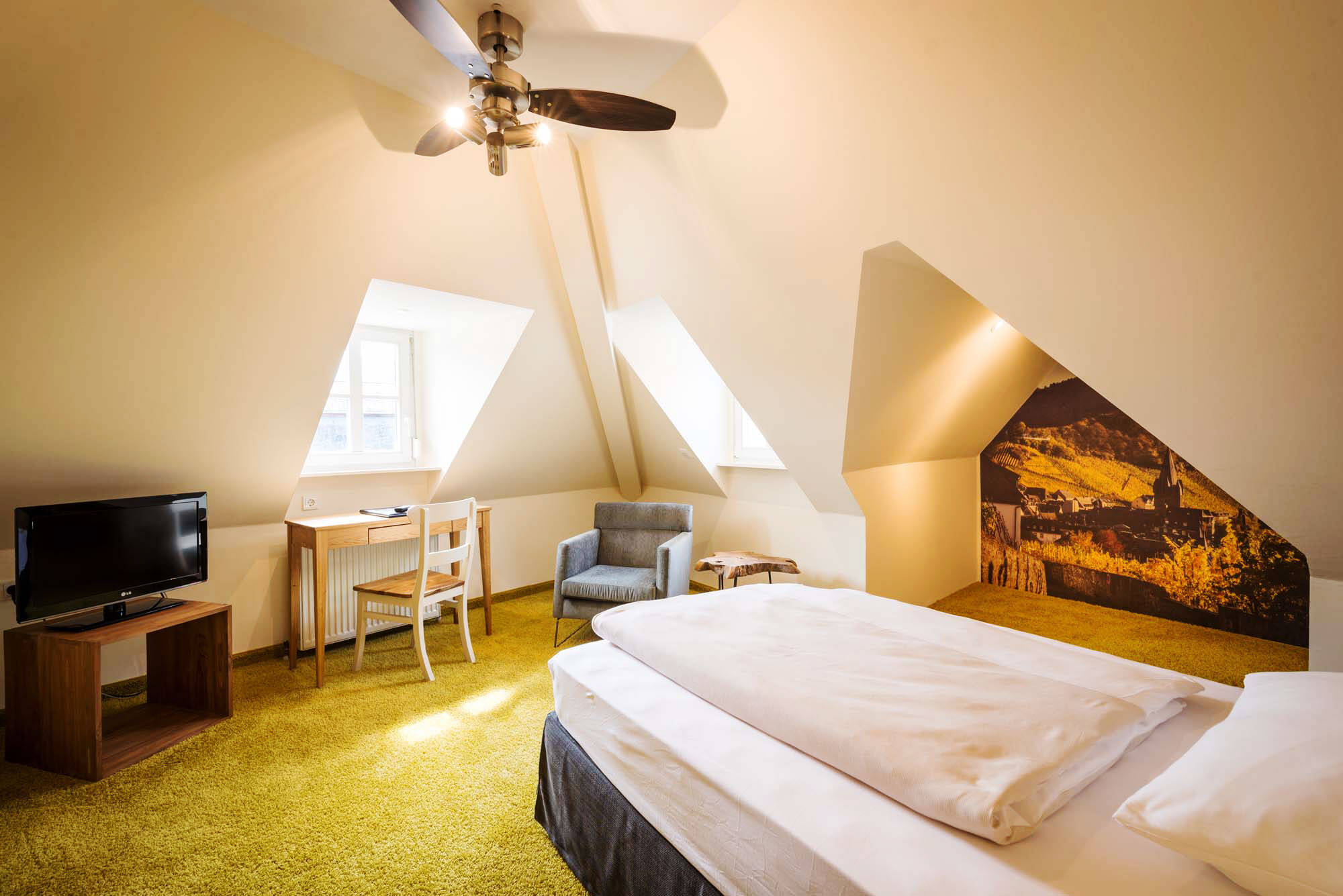 Ein Einzelbett steht in einem hellen Raum mit verwinkelter Decke. Der Teppichboden ist hellgrün. In der Ecke stehen ein kleiner Schreibtisch und ein grauer Sessel. Ein Erker bildet eine gemütliche Ecke, an dessen Wand ein großes Foto der Mosel hängt.