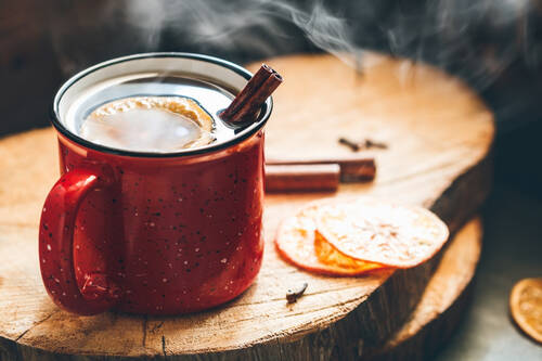 Eine dampfende Tasse Glühwein steht auf einem Holzbrett. Daneben liegen getrocknete Orangenscheiben und Zimtstangen als weihnachtliche Dekoration.