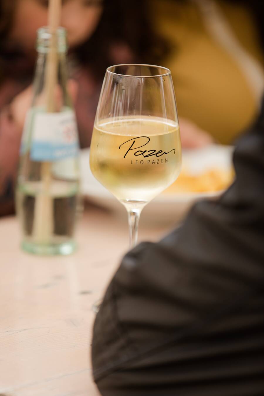 Ein Weinglas mit Weißwein steht auf einem Holztisch einer Bierzeltgarnitur. Im unscharfen Vorder- und Hintergrund sitzen Menschen auf den Bänken bei einem Fest.