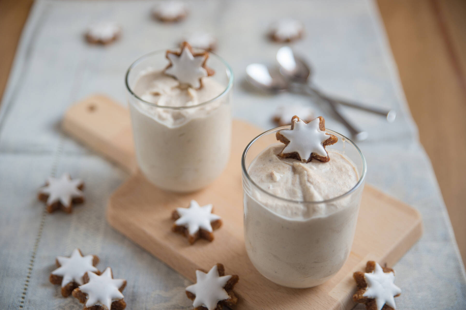 Auf einem Holzbrett stehen zwei Gläser, gefüllt mit einer Dessertcreme. Zimtsterne sind sowohl auf dem Dessert als auch auf dem Brett und Tisch verteilt. Im Hintergrund sind unscharf zwei Löffel zu erkennen.