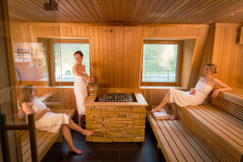 Drei Frauen in weißen Handtüchern befinden sich in der Sauna. Eine Frau sitzt rechts, eine links, und eine andere hält den Holzkübel in der Hand und gießt Wasser auf die Saunasteine für den Aufguss.