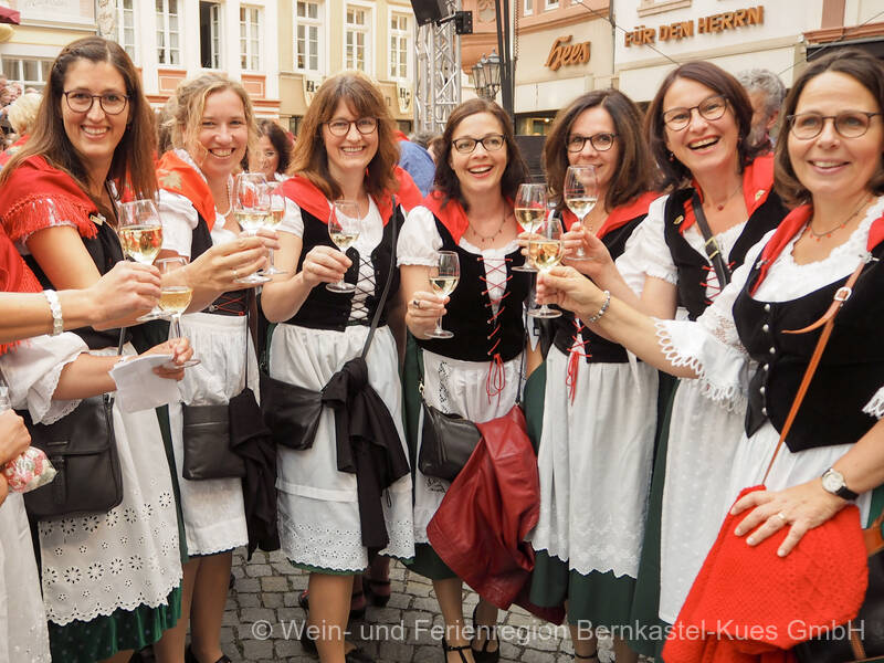 Sieben Frauen stehen nebeneinander im Moselblümchen-Outfit, stoßen mit ihren Weingläsern an und lachen in die Kamera.