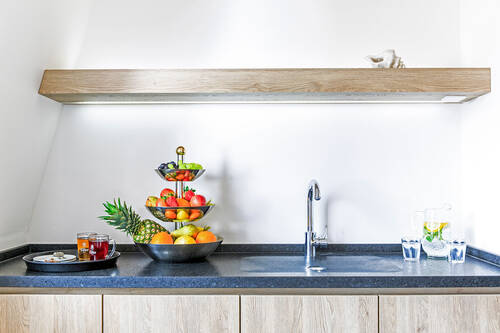 Eine Getränkeecke im Wellnessbereich des Deutschherrenhofs. Auf der linken Seite der Küchenzeile steht ein Tablett mit zwei Tees, daneben eine Etagere mit frischem Obst. Neben dem Waschbecken stehen eine Karaffe mit Wasser und zwei Gläser.