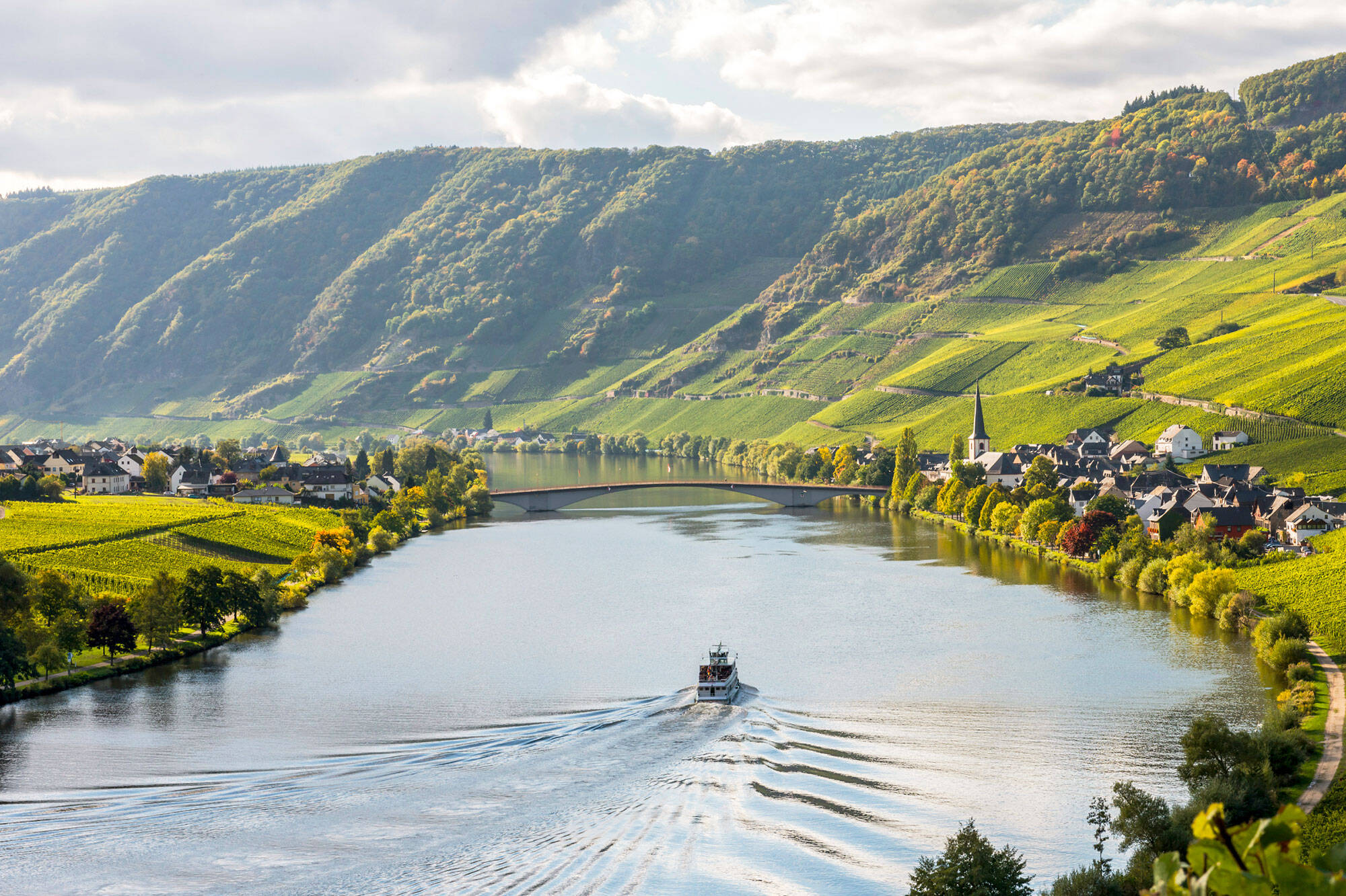 Ein Schiff fährt in der Mitte der Mosel. Hinter dem Schiff bilden sich Wellen auf dem Fluss. Auf beiden Seiten des Flusses sind Dörfer zu sehen, die durch eine Brücke miteinander verbunden sind. In der Ferne ragen steile Berge und Weinberge in den Himmel.