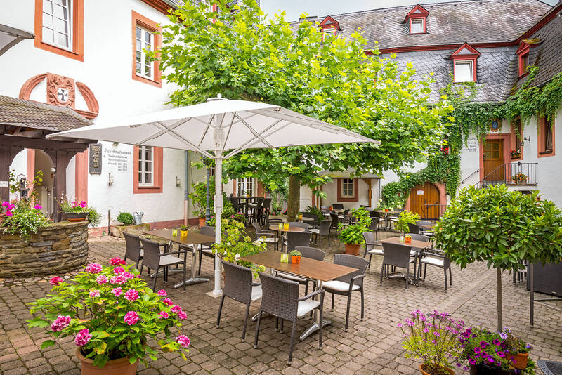 Im Innenhof des Hotels stehen mehrere Tische mit Stühlen unter einem großen Sonnenschirm. Ein blühender Baum und farbenfrohe Pflanzen sorgen für eine lebendige und einladende Atmosphäre.