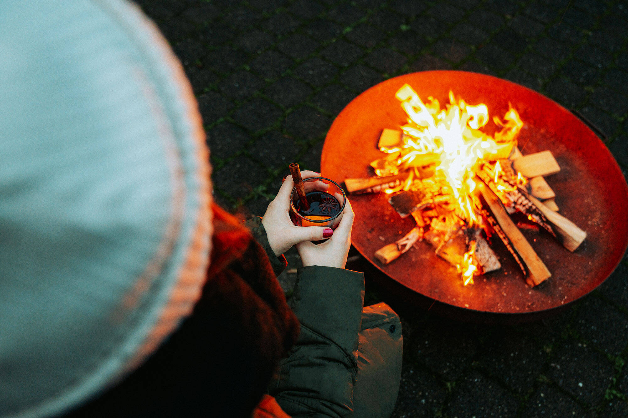 Eine Frau mit Mütze sitzt an einer Feuerschale und hält eine dampfende Tasse Glühwein in der Hand.