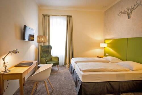 In einem kleinen, gemütlichen Doppelzimmer steht links ein Holzschreibtisch mit Stuhl unter einem Wandfernseher. In der Ecke steht ein großer grüner Sessel vor einem Fenster mit bodentiefen Vorhängen. Das Doppelbett hat ein knallig grünes Kopfteil, das durch das silberne Geweih an der Wand einen besonderen Akzent im Raum setzt.