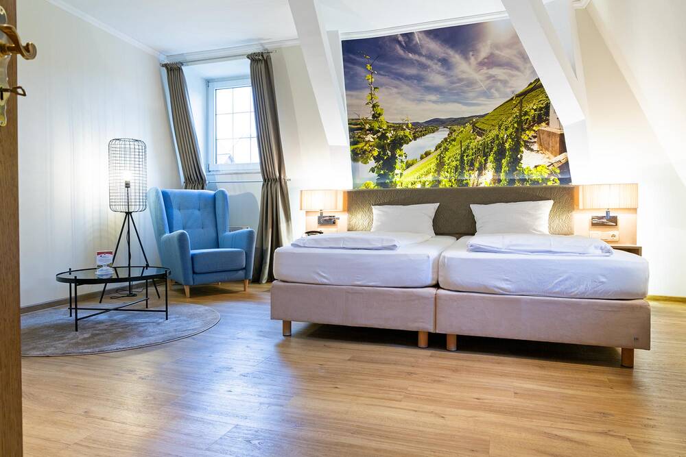 In einem modern gestalteten Zimmer steht ein beige-weißes Doppelbett auf hellem Holzboden. Hinter dem Bett erstreckt sich ein großes Wandbild mit Weinbergen und der Moselschleife. Links daneben stehen ein blauer Sessel, eine Leselampe und ein kleiner Tisch.