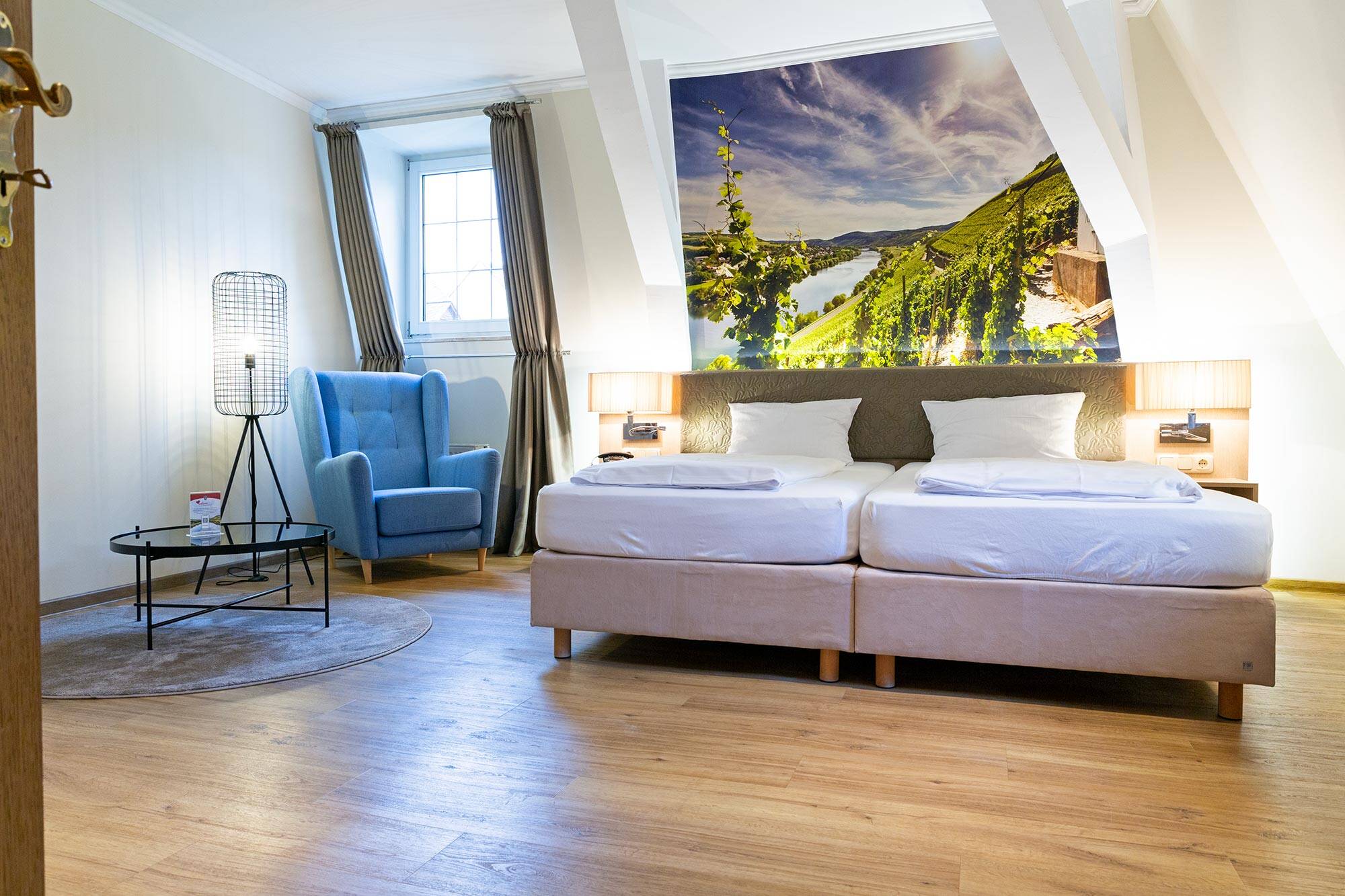 In einem modern gestalteten Zimmer steht ein beige-weißes Doppelbett auf hellem Holzboden. Hinter dem Bett erstreckt sich ein großes Wandbild mit Weinbergen und der Moselschleife. Links daneben stehen ein blauer Sessel, eine Leselampe und ein kleiner Tisch.