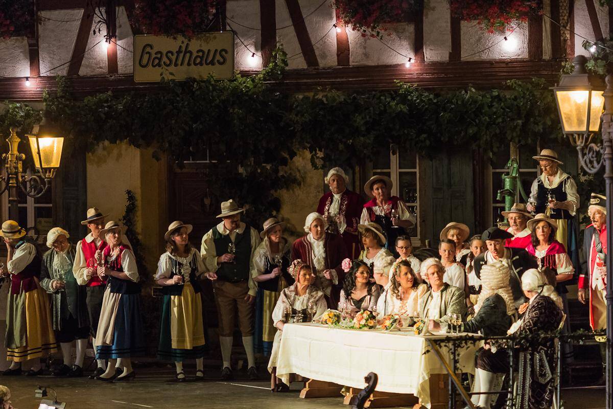 Eine Szene aus der Moseloperette. Viele Menschen stehen auf der Bühne um einen Tisch herum, an dem weitere Schauspieler sitzen.