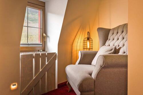 Gemütliche Sitzecke in einem Rieslingzimmer im Deutschherrenhof. Ein beiger Sessel steht in einem kleinen Erker. Vor dem Fenster ist ein Holzelement angebracht. Eine Lampe sorgt für gemütliches Licht.