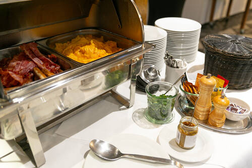 Auf dem Frühstücksbuffet steht ein Chafing Dish, das Rührei, Bacon und Würstchen warm hält. Daneben befinden sich Teller, Salz, Pfeffer, Senf und Lauchzwiebeln.