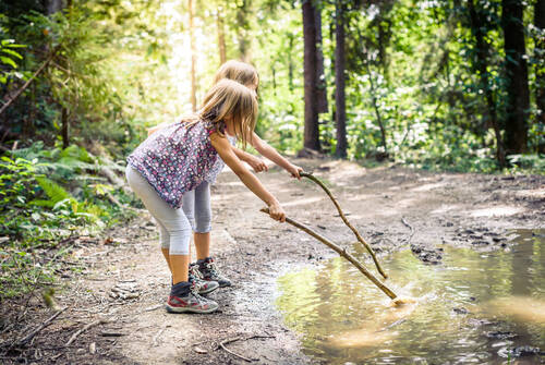 Zwei Kinder stehen in einem Waldstück neben einer großen Pfütze. Beide halten einen großen Stock und spielen damit in der Pfütze.