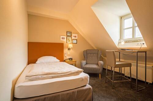 Helles, beiges Einzelzimmer im Deutschherrenhof. Am Fenster steht ein Schreibtisch, daneben ein grauer Sessel in der Ecke. Ein Einzelbett mit orangem Kopfteil steht im Raum. Über dem Nachttisch hängen mehrere Bilderrahmen mit Fotos.
