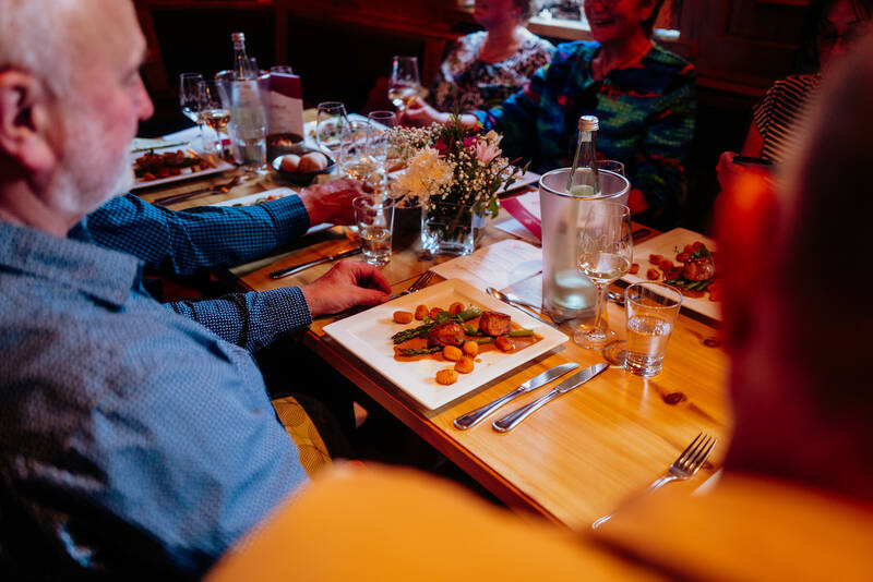 Mehrere Menschen sitzen gemeinsam an einem Tisch und essen. Vor ihnen stehen quadratische Teller mit einer Hauptspeise, die Spargel enthält. Ein Mann im Vordergrund hält ein Weinglas in der Hand und blickt auf sein Essen.