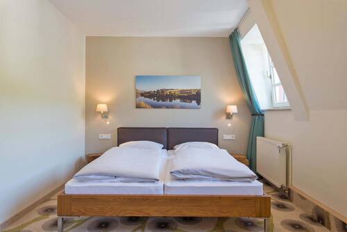 In einem hellgrün gestrichenen Raum steht ein Doppelbett aus Holz mit braunem Kopfteil auf einem gemusterten Teppich. Über dem Bett hängt ein Foto der Mosel. Der Vorhang am Fenster ist in einem auffälligem Petrolton gehalten.