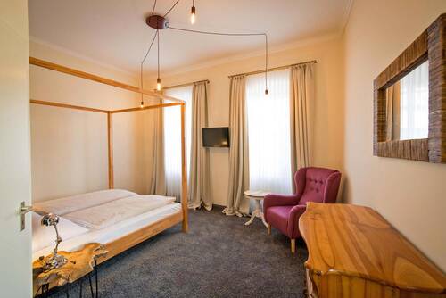 Ein helles, beige gestaltetes Doppelzimmer im Deutschherrenhof. Der dunkle Teppichboden und die hellen Holzmöbel ergänzen sich harmonisch. Als Highlight gibt es ein Himmelbett und einen lilafarbenen Sessel im Raum.