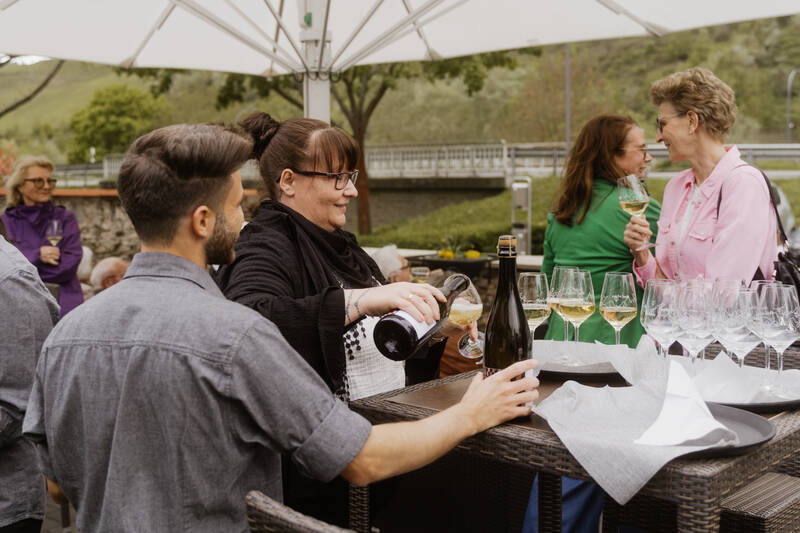 Zwei Mitarbeiter des Deutschherrenhofs schenken auf der Terrasse Sekt in Sektfläser auf dem Stehtisch aus. Im Hintergrund sieht man Gäste, die sich unterhalten.