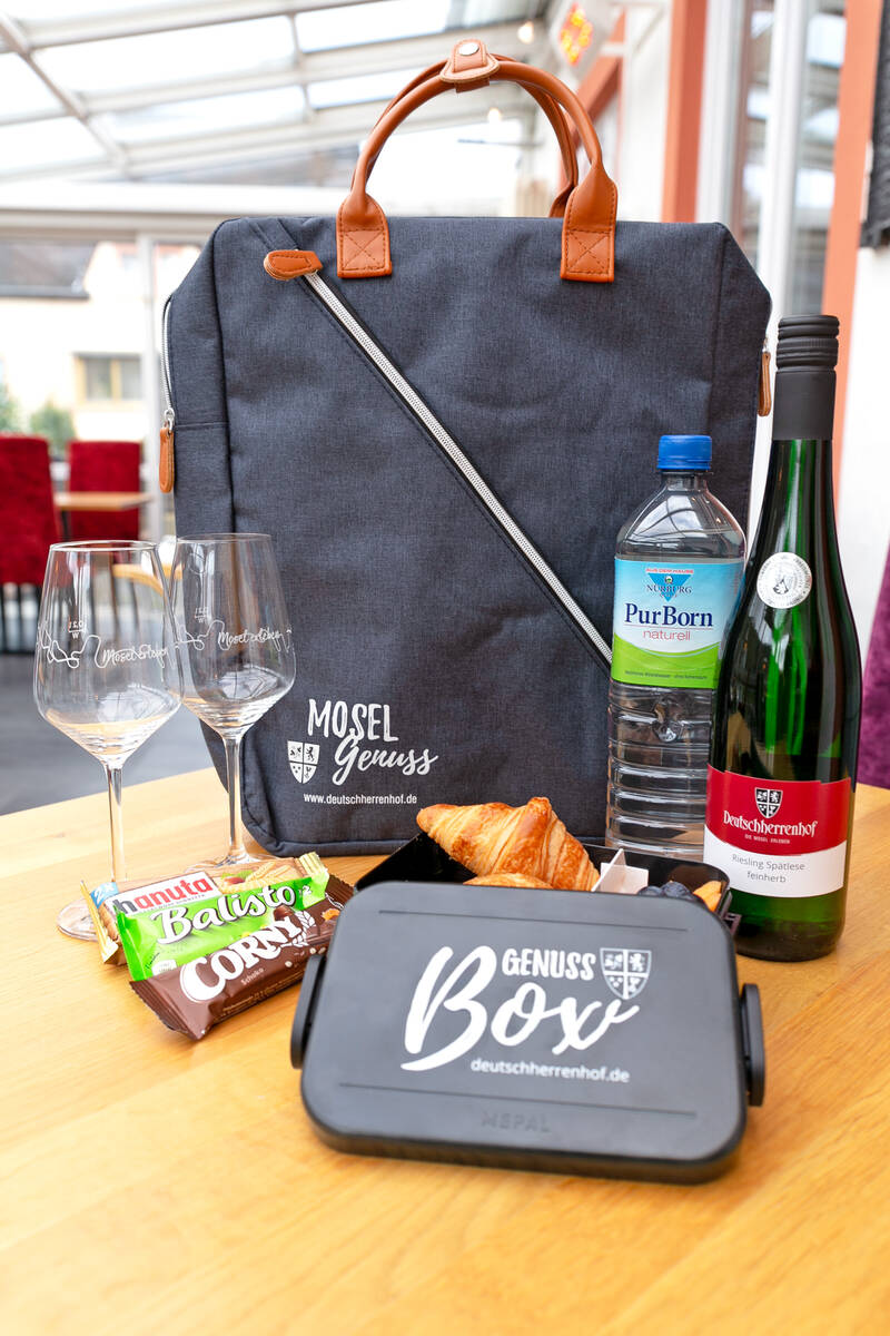 Auf einem Tisch steht ein Rucksack mit der Aufschrift ‚Moselgenuss‘. Davor befinden sich zwei Weingläser, drei verschiedene Müsliriegel, eine Weinflasche, eine Wasserflasche und eine halbgeöffnete Genussbox, aus der ein Croissant hervorblitzt.