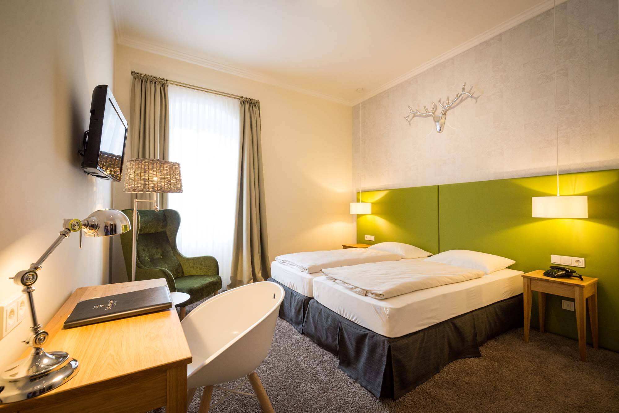 In einem kleinen, gemütlichen Doppelzimmer steht links ein Holzschreibtisch mit Stuhl unter einem Wandfernseher. In der Ecke steht ein großer grüner Sessel vor einem Fenster mit bodentiefen Vorhängen. Das Doppelbett hat ein knallig grünes Kopfteil, das durch das silberne Geweih an der Wand einen besonderen Akzent im Raum setzt.