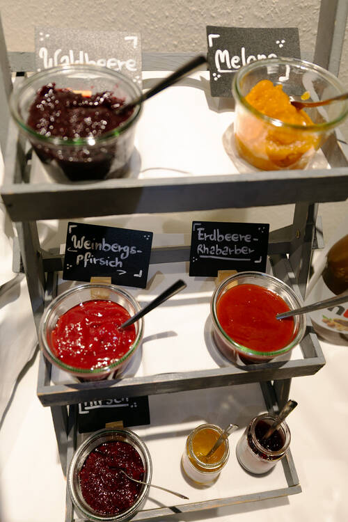 Eine Etagere aus Holz zeigt eine Auswahl hausgemachter Marmeladen. Auf den drei Etagen sind sieben verschiedene Gläser platziert, jedes mit einem Löffel darin. Hinter jedem Glas befindet sich eine kleine Schieferplatte mit der Bezeichnung der jeweiligen Marmeladensorte.