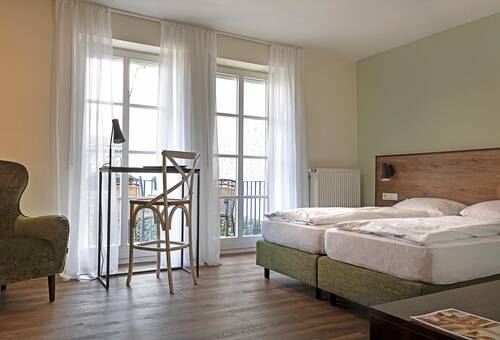 Das Doppelzimmer im Gästehaus ist in hellen Grün- und Brauntönen gestaltet. Das Doppelbett mit Holz-Kopfteil steht auf hellem Holzboden. Links im Bild befindet sich ein gemütlicher Sessel, daneben ein Schreibtisch mit rustikalem Holzstuhl. Zwei Balkontüren, eingerahmt von weißen Vorhängen, bieten einen Blick auf den Balkon.